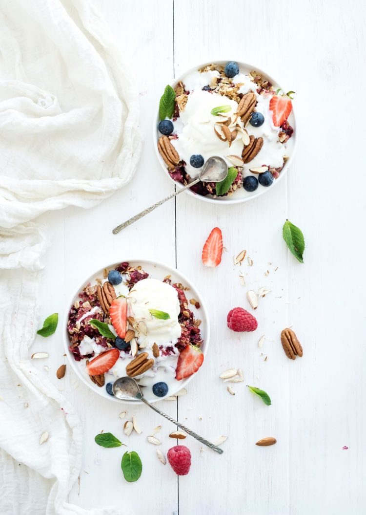 Healthy breakfast. Berry crumble with fresh blueberries, raspberries, strawberries, almond, walnuts, pecans, yogurt, and mint in ceramic plates over white wooden surface, top view